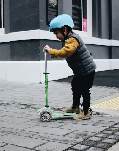 boy riding a green mini deluxe 3 wheel scooter on footpath